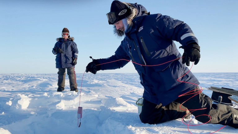 Seismic sampling involved drilling a small hole into the ice and then placing a small, concentrated explosive (PETN) into the ice. When triggered, sound vibrations are sent into the ice and the echo timing, strength and direction provide information on ice thickness and the materials beneath. The image shows an explosive being lowered into the hole by the geophysical team